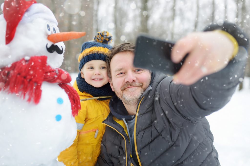Little boy and his father taking selfie on background of snowman in snowy park. Active outdoors leisure with children in winter.
