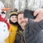 Little boy and his father taking selfie on background of snowman in snowy park. Active outdoors leisure with children in winter.